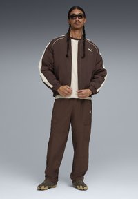 Veste aviateur marron avec des accents blancs, superposée à un haut crème, assortie à un pantalon cargo marron et des baskets vert olive, sur un fond gris.