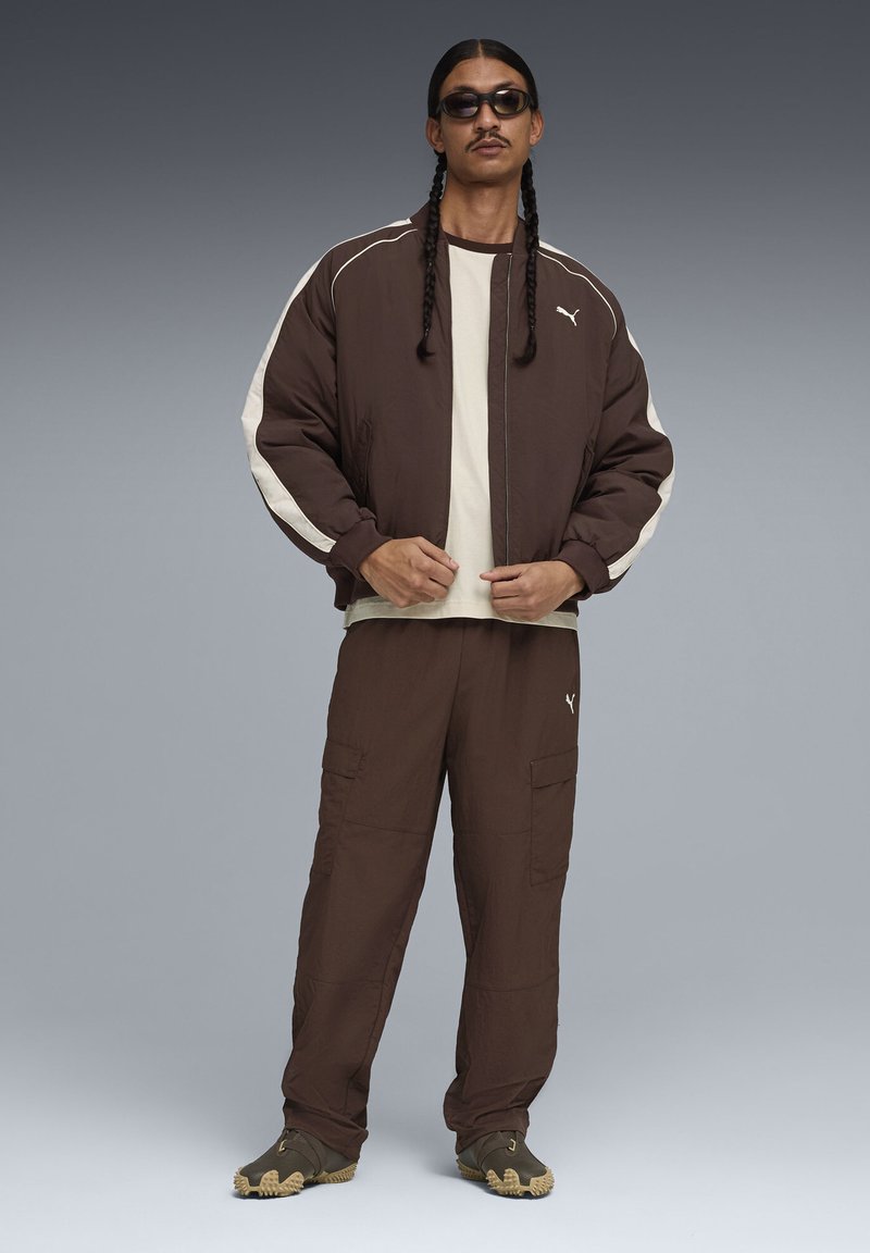 Veste aviateur marron avec des accents blancs, superposée à un haut crème, assortie à un pantalon cargo marron et des baskets vert olive, sur un fond gris.