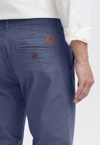 Un chino bleu marine dispose d'une poche arrière avec fermeture à bouton et d'un patch de marque en cuir marron. Le tissu est lisse, avec une coupe soignée.