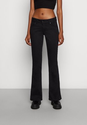 Woman wearing black flared jeans, black crop top, and black shoes, standing against a plain light gray background.