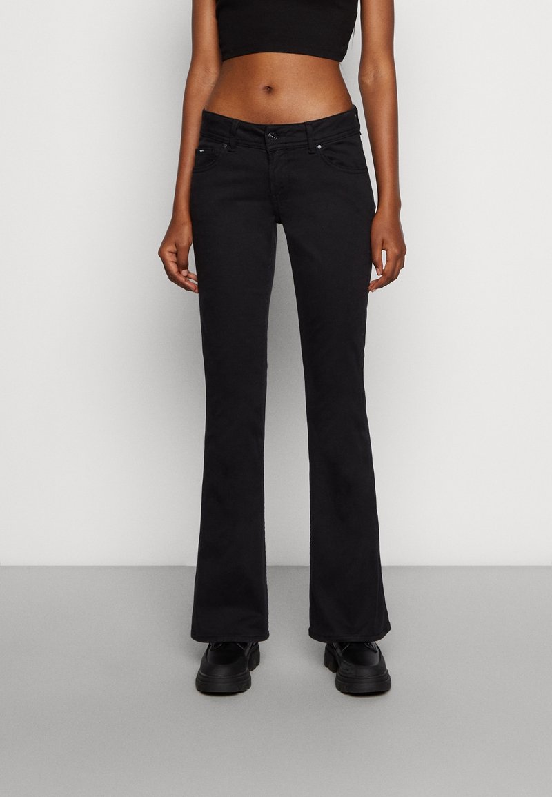 Femme portant un jean évasé noir, un crop top noir et des chaussures noires, debout devant un fond gris clair uni.