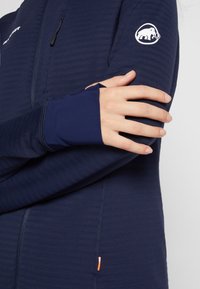 Dunkelblaue Fleecejacke mit gerippter Textur, Reißverschluss, Daumenlöchern und kontrastierendem orangefarbenen Akzent am Saum. Logo am Ärmel.