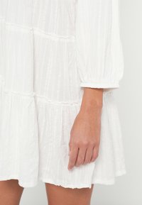 Weißes, strukturiertes Kleid mit langen Puffärmeln und einem fließenden, gestuften Design. Der Stoff weist dezente vertikale Linien und geraffte Akzente auf.