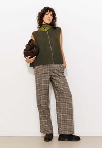Pull sans manches vert olive avec un zip sur le devant, associé à un pantalon large à carreaux marron. Bottines chelsea marron foncé, tenant un sac marron.