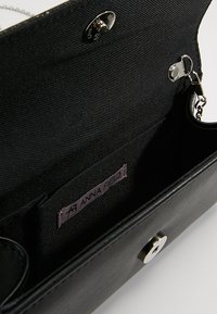 Portefeuille noir avec un tissu texturé et des accents lisses, comportant une fermeture à pression, un compartiment zippé et une étiquette violette portant la mention "ANNA FIELD."