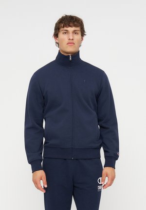 Jeune homme portant une veste zippée bleu marine et un pantalon de survêtement assorti, debout devant un fond clair uni.