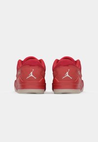 Raudoni zomšiniai sportbačiai su lygiu paviršiumi, turintys baltas Jumpman logotipų etiketes ant kulnų ir permatomą pilką padą. Minimalistinio dizaino akcentai.
