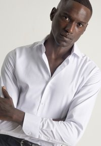 Camisa blanca de vestir con un diseño ajustado, ligero brillo y un cuello estructurado. Presenta un frente de botones y mangas largas con puños abotonados.