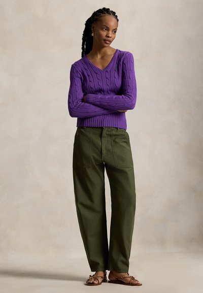 Μωβ πουλόβερ με πλέξη καλτσών και V-λαιμόκοψη, συνδυασμένο με χαλαρά ελιάς πράσινα παντελόνια. Καφέ σανδάλια με διακοσμητικά στοιχεία. Ουδέτερο φόντο.