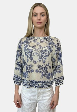 Giovane donna con capelli biondi lisci che indossa un maglione beige con intricati motivi floreali blu e pantaloni bianchi con dettagli testurizzati.