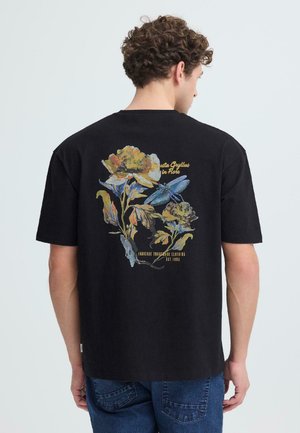 Homme portant un T-shirt noir avec un motif coloré de fleurs et une libellule bleue dans le dos, accompagné d'un jean bleu, dos tourné à la caméra.