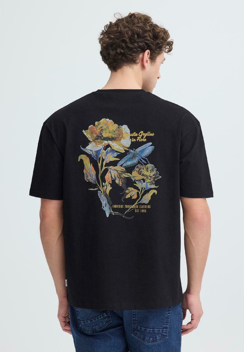 Άνδρας που φοράει μαύρο T-shirt με πολύχρωμο λουλούδι και μπλε λιβελούλα στο πίσω μέρος, συνδυασμένο με μπλε τζιν, με την πλάτη προς την κάμερα.