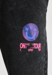 Svart denim-stoff med et flerfarget grafisk design med et smilende ansikt og stilisert tekst som leser "ON TOUR" i rosa og blå nyanser.