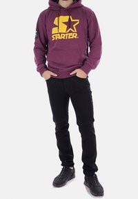 Sudadera granate con logo amarillo "STARTER", que incluye un bolsillo canguro, pantalones negros de corte ajustado y zapatillas negras.