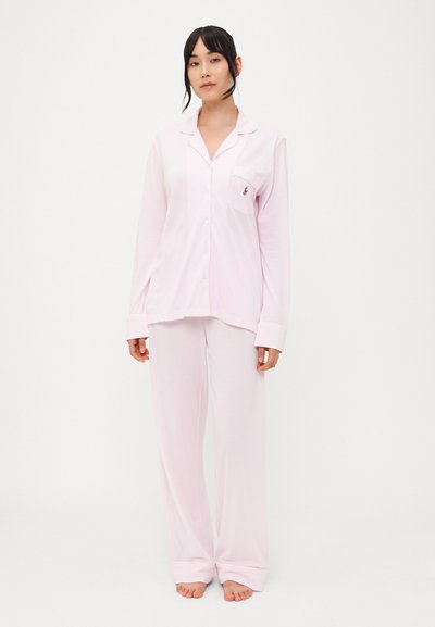 Ensemble de pyjama rose comprenant un haut à boutons avec une poche et des manches longues, associé à un pantalon ample. Les deux sont fabriqués en tissu doux.