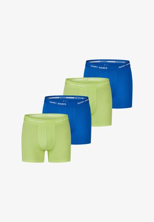 Limettengrüne Baumwollboxershorts mit einem weißen Bund, auf dem "HAPPY SHORTS" steht, getragen von einem Modell, das sich an einer Holzoberfläche lehnt.