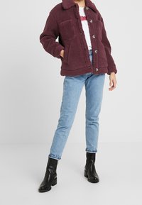 Persona vistiendo una chaqueta de sherpa color marrón, camiseta blanca, jeans de mezclilla azul claro y botas negras hasta el tobillo, de pie contra un fondo blanco liso.
