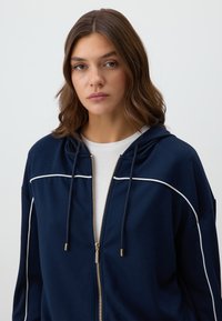 Navy zip-up hoodie with white piping details, drawstring hood, and soft fabric. Features a gold zipper and a relaxed fit.