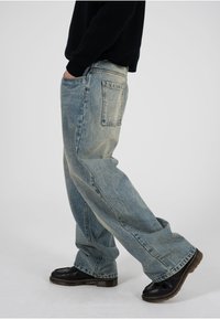 Jeans de mezclilla azul claro con un corte holgado, textura deslavada y diseño de cinco bolsillos, con un único bolsillo trasero. Combinados con zapatos negros.