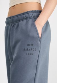 Joggers de mezcla de algodón grises con cintura elástica, bolsillo lateral y logo impreso "NEW BALANCE 1906" en el muslo izquierdo. Textura suave.