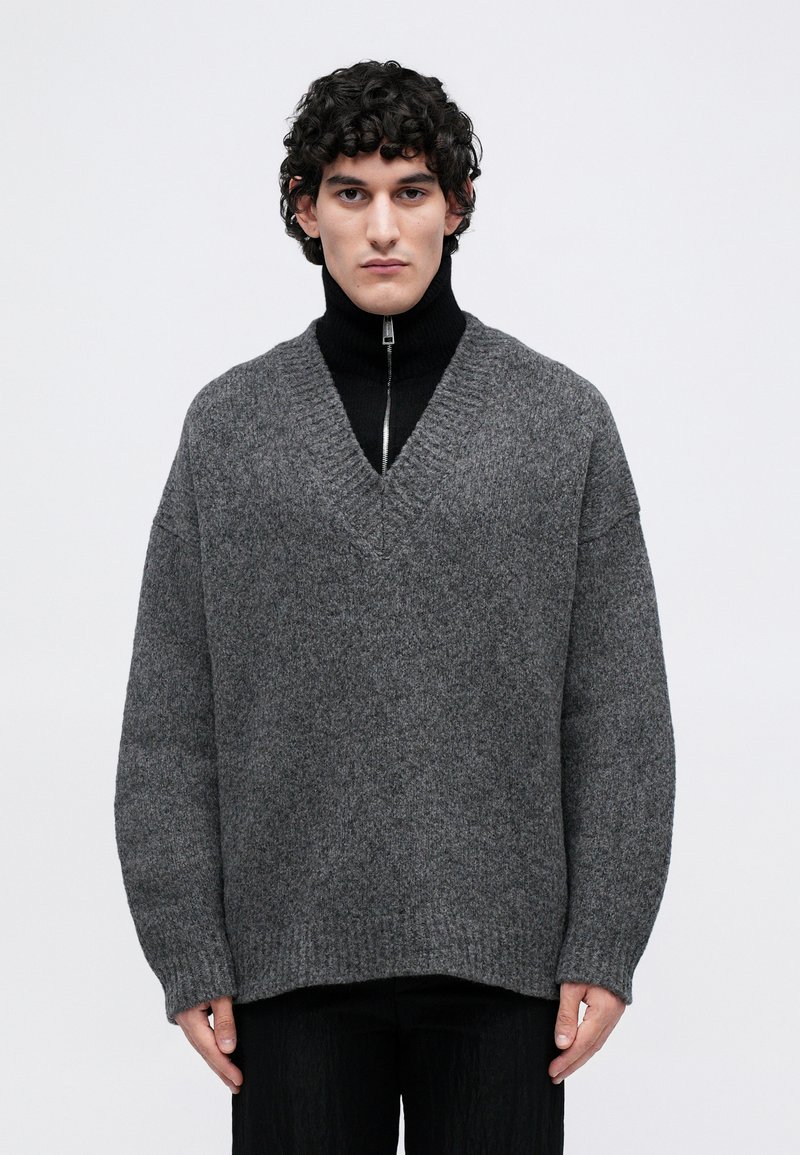 Mlad moški s kodrastimi lasmi, oblečen v sivo preveliko V-izrezano jopico preko črnega zip-up turtlenecka, stoji pred enostavno belo ozadje.
