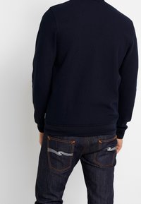 Man som bär en marinblå långärmad tröja och mörkblå denimjeans med orange sömmar och ett vitt vågmönster på bakfickorna, vänd bort.