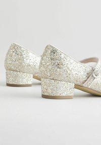 Glitzernde silberne Low-Heels mit dicker Absatzzahl, runder Zehenpartie und dekorativem Riemen mit einem runden Hardware-Element.