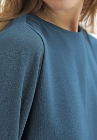 Teksturowana niebieska bluzka z dekoltem w łódkę i krótkimi rękawami, z delikatnym wzorem splotu, który dodaje wizualnego zainteresowania.