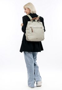 Beige Stoff-Rucksack mit einem oberen Griff, verstellbaren Trägern, einer vorderen Reißverschlusstasche und einem Lederakzent am Reißverschlussgriff.
