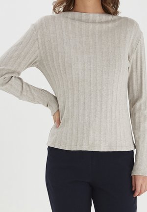 Žena v světle šedém žebrovaném tričku s dlouhými rukávy a tmavě modrých kalhotách, s jednou rukou na kyčli, stojící před bílým pozadím.