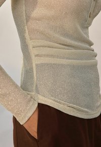 Lähikuva henkilöstä, jolla on päällä kiiltävä beige pitkähihainen paita, joka on pujotettu tummanruskeisiin housuihin, ja käsi taskussa.