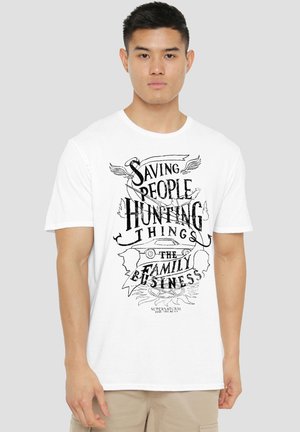 Young man wearing a white T-shirt with black text and graphics reading "Saving People Hunting Things The Family Business."