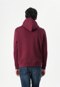 Burgunderfarget hettegenser laget av mykt stoff, med avslappet passform, lange ermer og ribbestrikkede mansjetter og kant. Brukt med blå jeans.