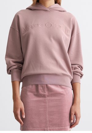 Frau trägt einen staubrosa Marc O’Polo Hoodie mit hochgekrempelten Ärmeln und einen passenden, gerippten Cordrock.