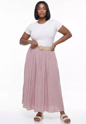 Kobieta o prostych czarnych włosach, ubrana w biały krótki top, beżowy pasek, długą różową spódnicę i beżowe sandały, stojąca z jedną ręką na biodrze.