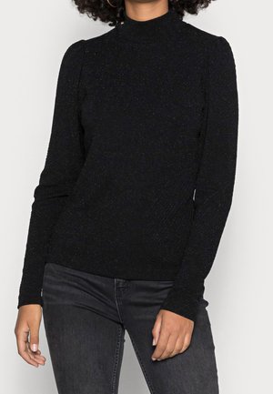 Femme portant un pull à col roulé noir texturé à manches longues et un jean gris foncé, debout devant un fond clair uni.