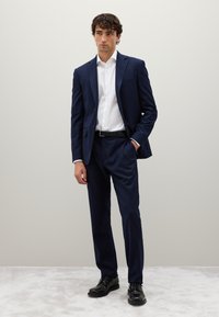 Traje navy con un patrón a cuadros, camisa blanca, cinturón negro y zapatos de vestir negros. La tela parece suave y ligeramente estructurada.