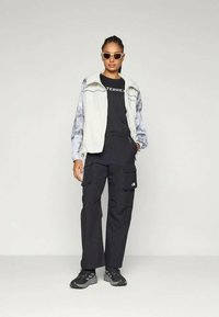 Gilet gris clair à motifs, t-shirt noir avec logo, pantalon cargo noir et baskets noires ; des lunettes de soleil complètent la tenue.