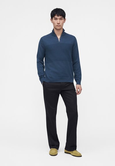 Homme debout, mains dans les poches, portant un pull bleu à fermeture éclair quart, pantalon noir et mocassins jaunes sur fond blanc.