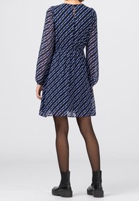 Námořnické šaty s geometrickým modrým vzorem, průhlednými dlouhými rukávy, elastickým pasem a detailem klíčové dírky na zádech. Nosené s černými kotníkovými botami.