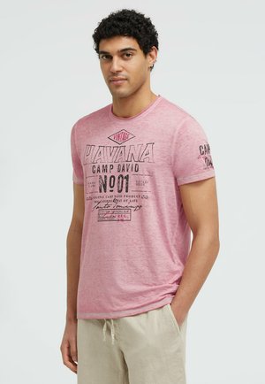 Junger Mann mit lockigem schwarzen Haar trägt ein pinkes T-Shirt mit dem Text "Havana Camp David No01" und hellbeigen Hosen, steht mit einer Hand in der Tasche.