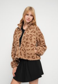 Μπεζ τζάκετ από συνθετική γούνα με σχέδιο λεοπάρ, ψηλό γιακά, κλείσιμο με φερμουάρ και πλαϊνές τσέπες. Φορεμένο πάνω από μαύρο τοπ και φούστα.