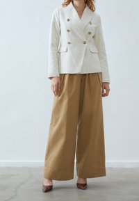 Krémový dvouřadý blazer s texturovanou tkaninou, zlatými knoflíky, v kombinaci s širokými hnědými kalhotami a hnědými vzorovanými podpatky.