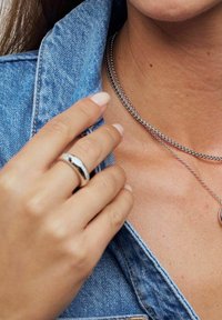 Silver ring på ringfingret, lager på lager av silverkedjor, denimjackans krage, jämn hudtextur och nagellack i neutrala toner.