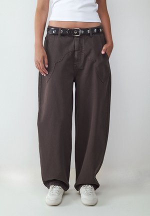 Persona que lleva puestos pantalones anchos marrón oscuro con grandes bolsillos delanteros, un cinturón negro con tachuelas, zapatillas blancas y un top blanco corto, de pie con las manos en los bolsillos.