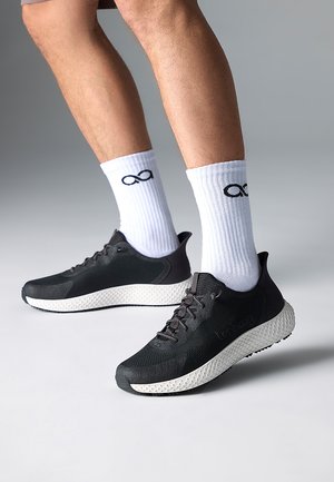 Beine mit weißen mittellangen Socken, die schwarze Unendlichkeitssymbole tragen, und schwarzen Sportschuhen mit strukturierten weißen Sohlen auf einem hellgrauen Boden.