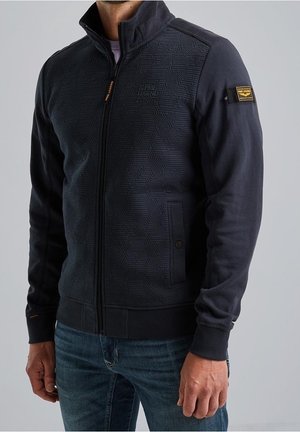 Man wearing a navy blue zip-up jacket with textured geometric pattern and patches, paired with blue jeans, standing against a gray background.