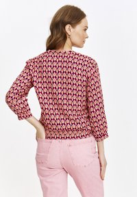 Gemusterte Bluse in Rottönen mit Augenschablonen, gerafften Schultern und langen Ärmeln; getragen mit hellrosa Hosen, von hinten dargestellt.