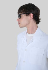 Camisa blanca de manga corta con dos bolsillos frontales y un collar; llevada sobre una camiseta de cuello redondo de color gris claro. Gafas de sol negras completan el atuendo.