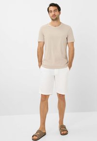Beige kortærmet t-shirt, hvide shorts og brune klip-klapper. T-shirten har en rund halsudskæring og en glat tekstur; shortsene er knælange.